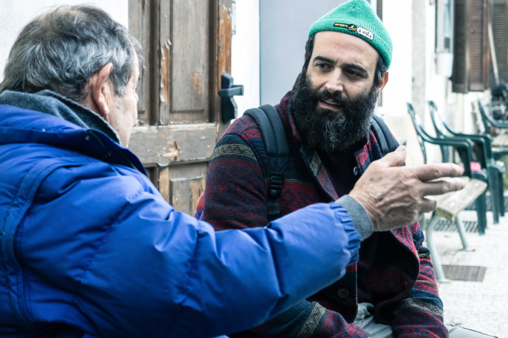 Un uomo con la barba e il cappello verde, parla con un signore più anziano dalla giacca blu. Sono seduti su una panchina.