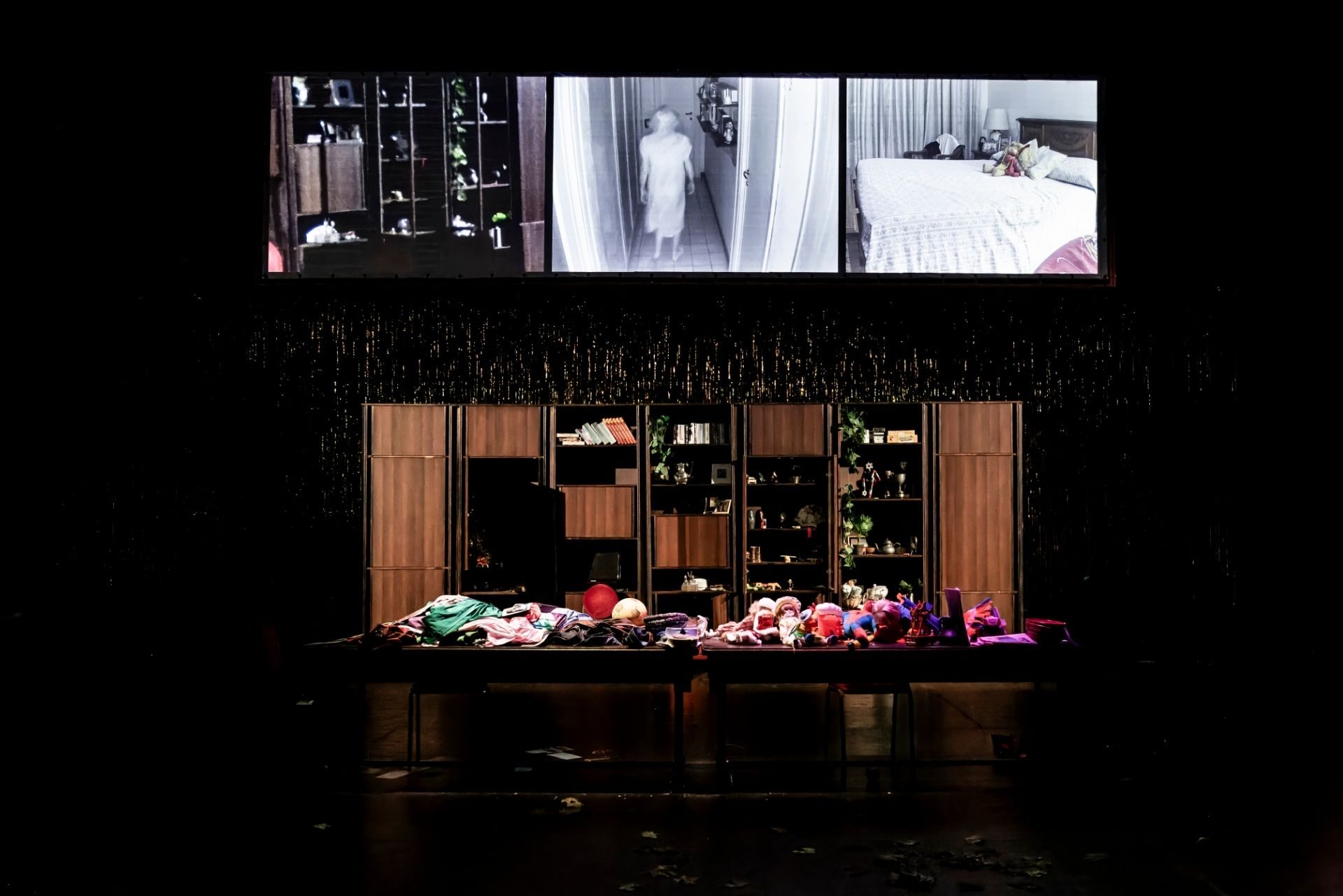 La foto mostra la scenografia di uno spettacolo su palco. Sul palco, due tavoli pieni di oggetti indefiniti. Sopra la scena uno schermo con proiezioni video.
