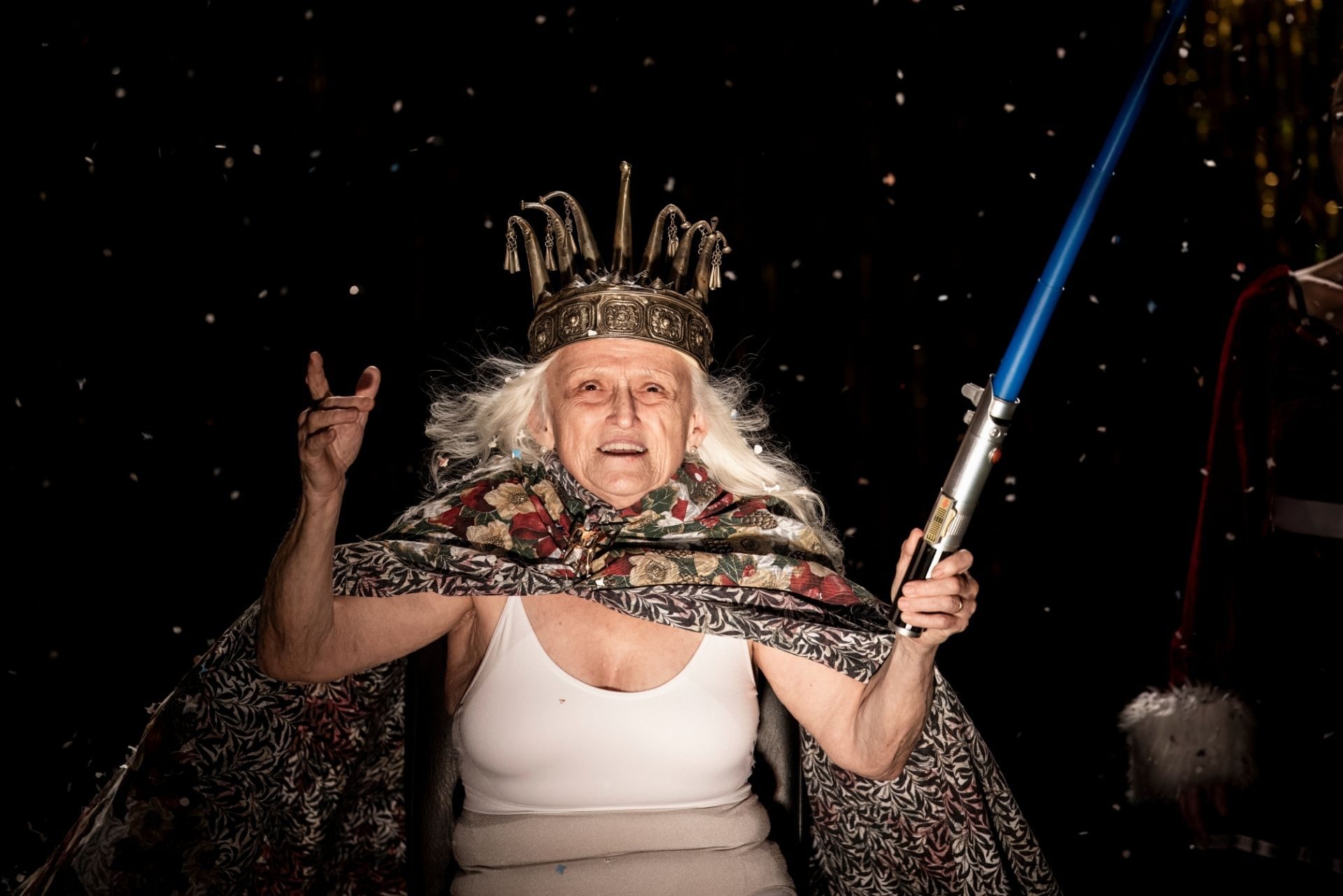 Un'anziana signora guarda verso la luce. Ha lunghi capelli bianchi, indossa una corona e un mantello, tiene in mano una spada laser.