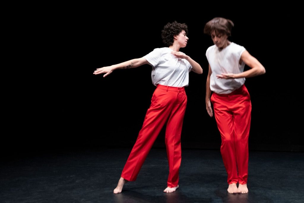 Due performer vestiti di bianco e rosso. Cristina Rizzo salta. Diana Anselmo tende un braccio.