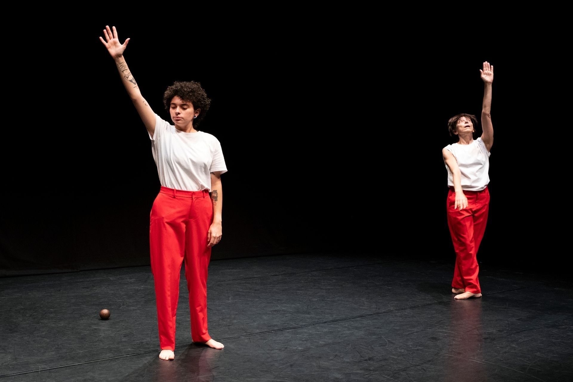 Due performer vestiti di bianco e rosso. Cristina Rizzo alza il braccio sinistro. Diana Anselmo tende il braccio destro.