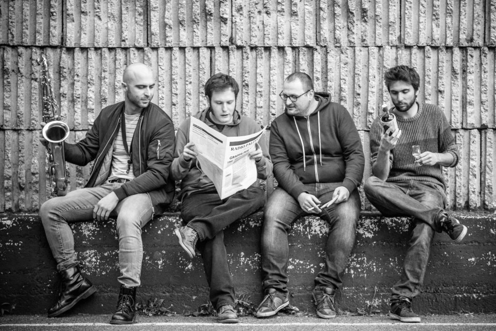 Quattro ragazzi sono seduti su un muretto: uno legge il giornale, uno fuma, uno beve del vino, uno regge un sassofono.