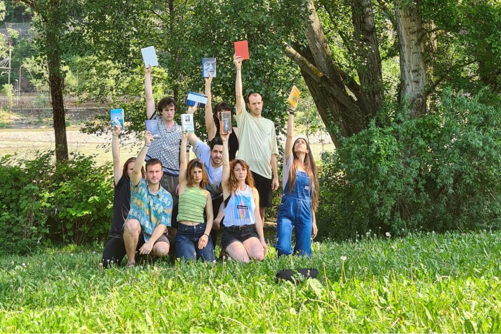 Un gruppo di ragazzi e ragazze in un prato. Tutti sollevano in aria un libro.
