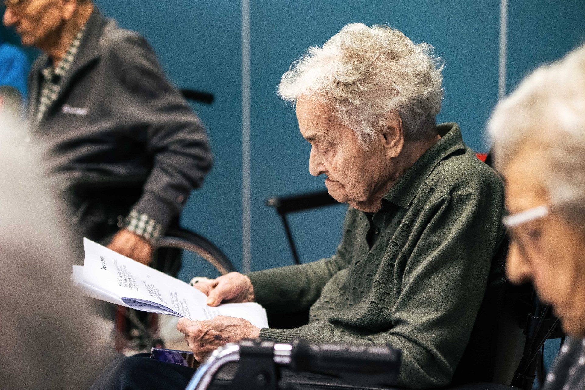 Un'anziana signora indossa un maglione verde scuro. Legge da alcuni fogli che tiene in mano.