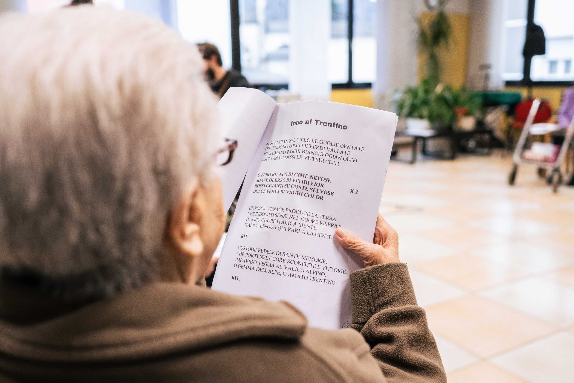 Un'anziana signora siede dandoci la schiena. Sta leggendo, su un foglio, l'Inno al Trentino
