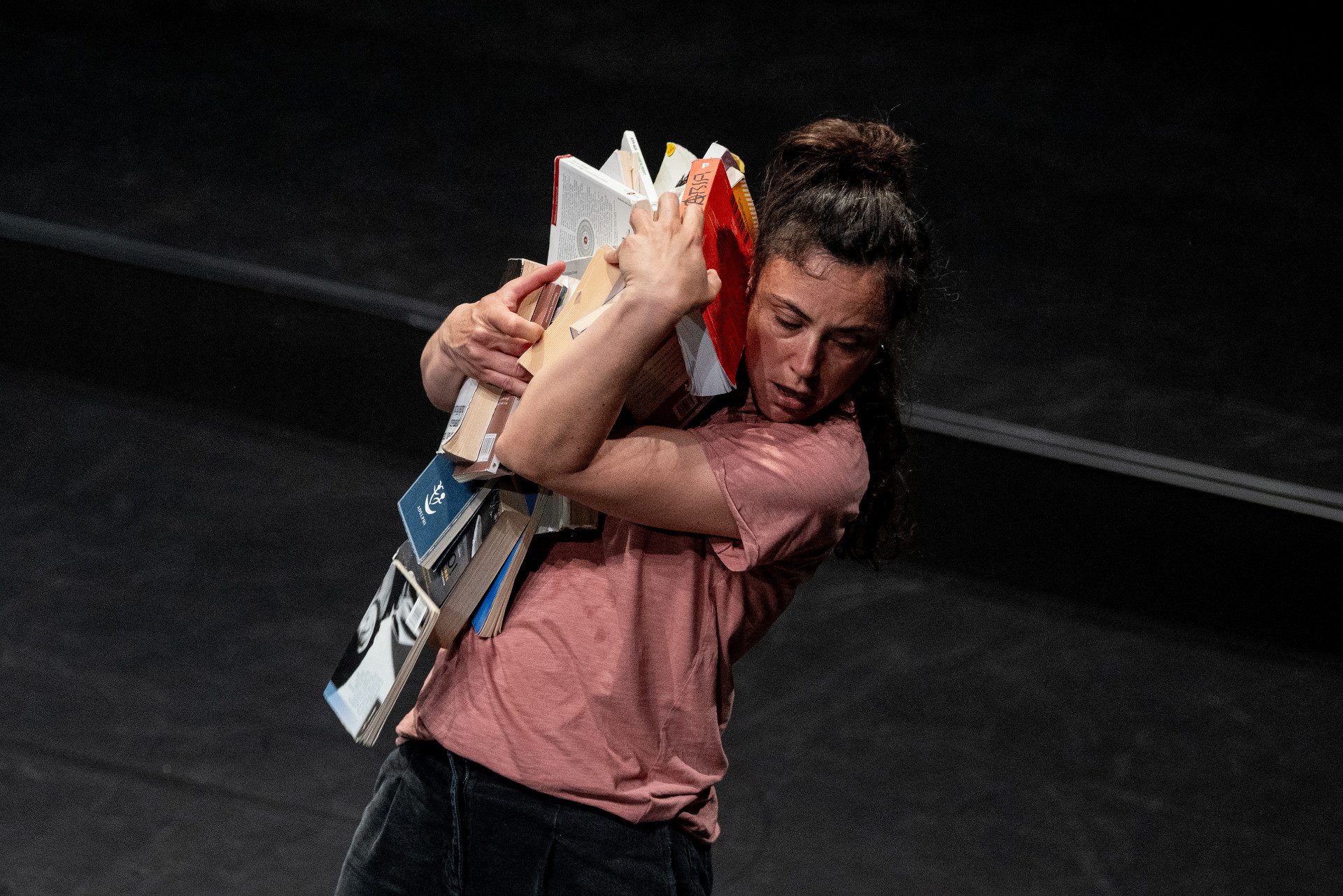 Una donna con capelli raccolti tiene stretti tra le braccia molti libri che sembra stiano per cadere.