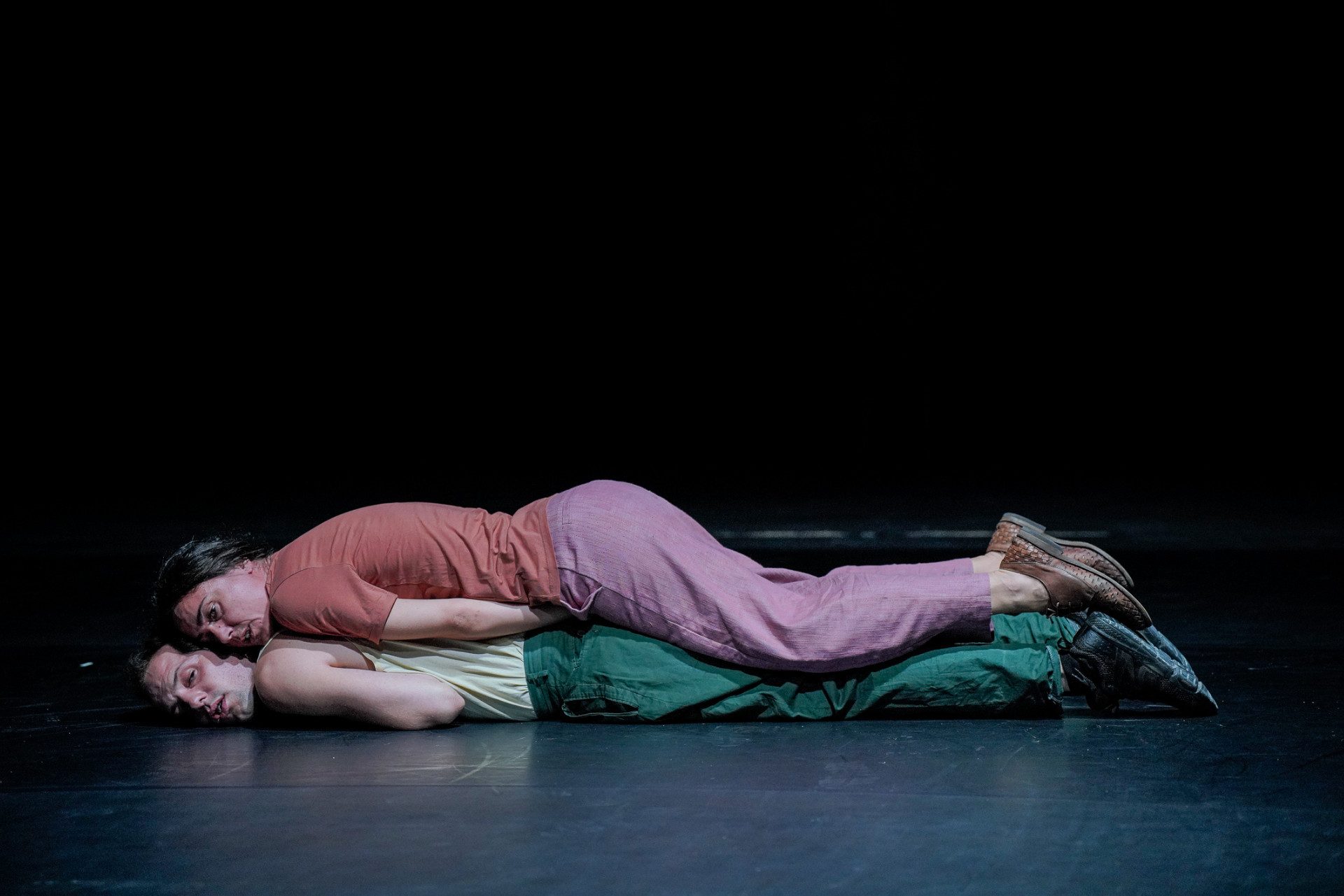 Un'attrice e un attore sono sdraiati sul palco una sopra l'altro. Sono vestiti.