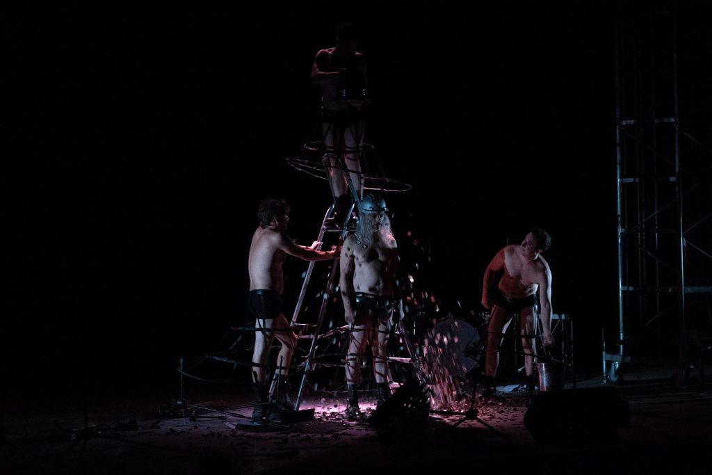 In ambientazione oscura, quattro uomini a petto nudo sono posizionati nei pressi di una scala a ventaglio.