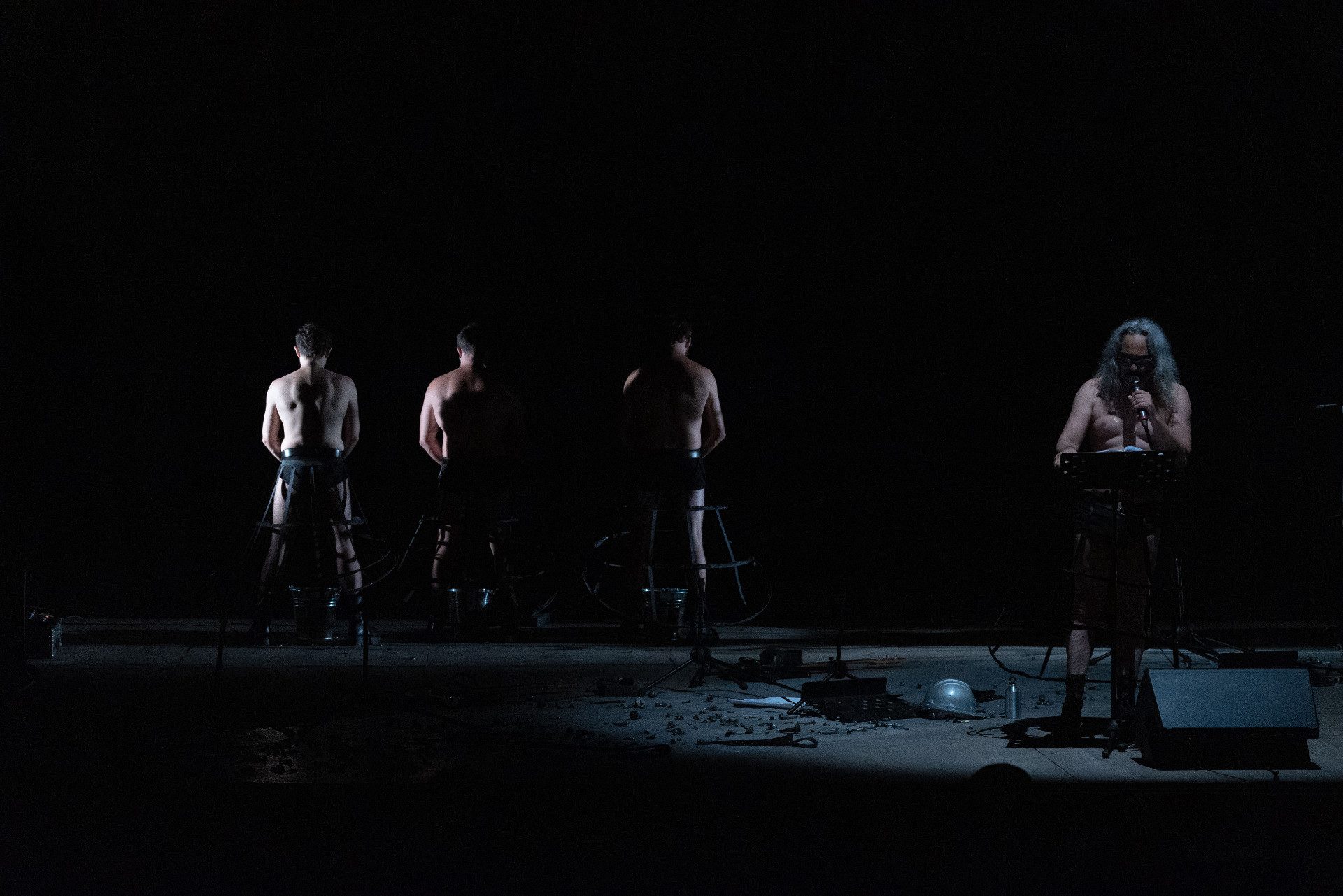 Quattro uomini a petto nudo: uno legge un testo al microfono. Tre ci voltano le spalle.