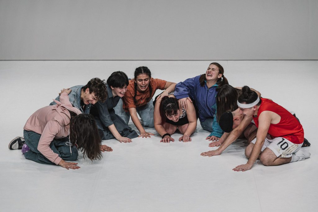 Nove attori adolescenti, seduti a terra in semicerchio, urlano e piangono.