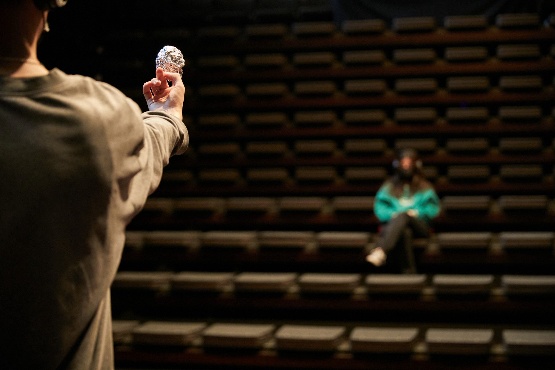 Una persona è seduta in una platea vuota. Guarda un'altra persona, che sta sul palco con in mano una pallina di stagnola.