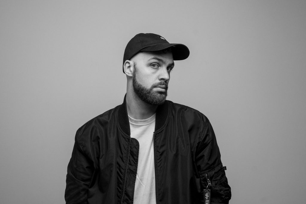 Il rapper Drimer. Indossa un cappellino con visiera nero, maglia bianca e giacca.