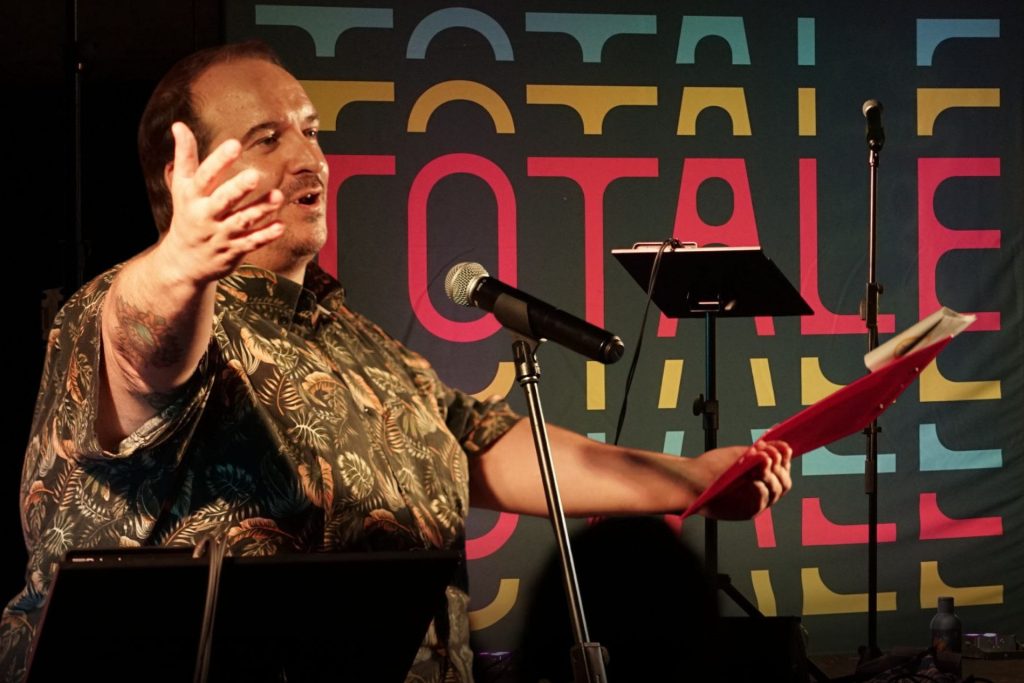 Jonathan Zenti tiene le braccia aperte e parla al microfono. Sullo sfondo la scritta TOTALE.