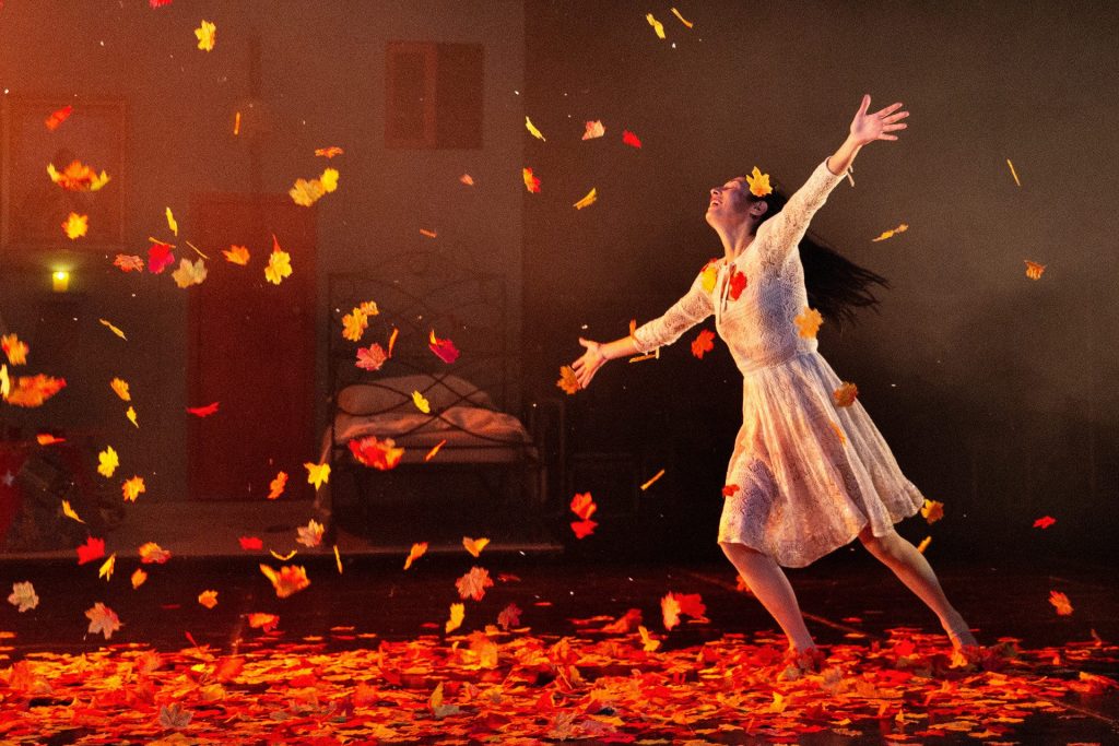Una donna vestita con un vestito bianco danza a braccia aperte mentre dall'alto cadono foglie rosse.