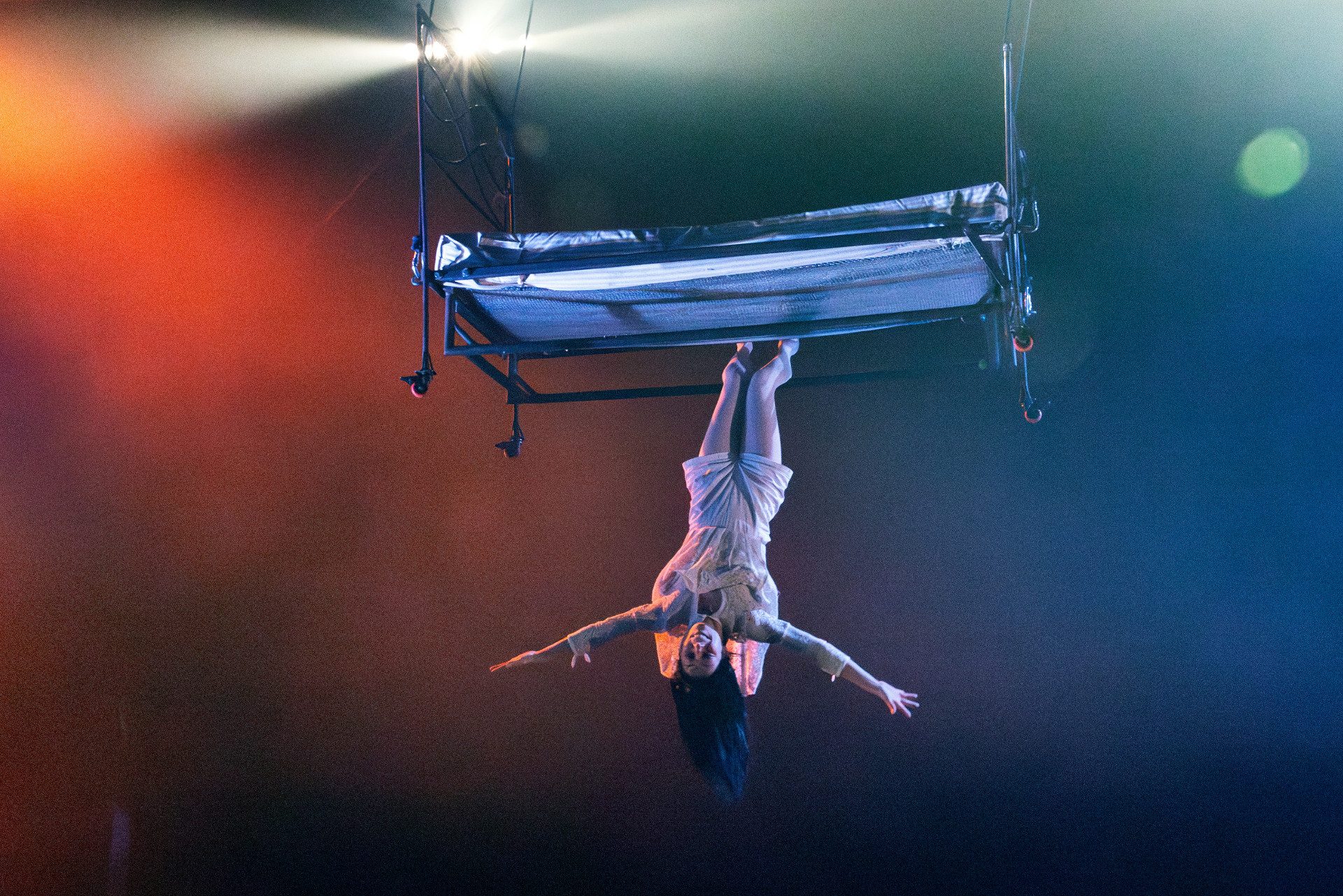 Una donna vestita con un lungo vestito bianco sta a testa in giù appesa con i piedi a un letto sollevato in aria.