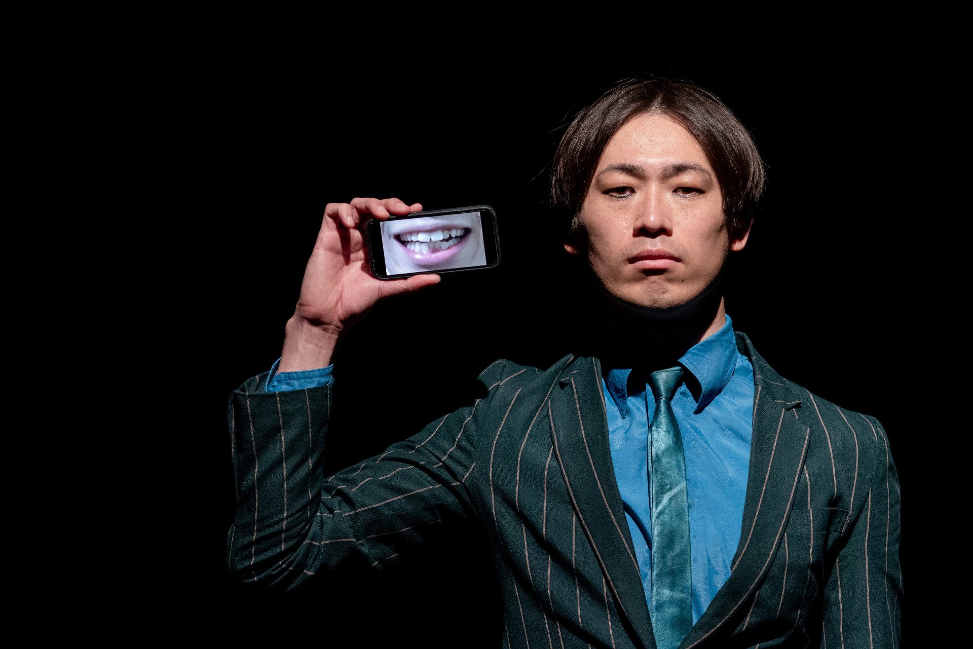 Il danzatore giapponese Kenji Shinohe ci guarda con espressione seria. Tiene alzato in mano il suo telefono che mostra una bocca sorridente.