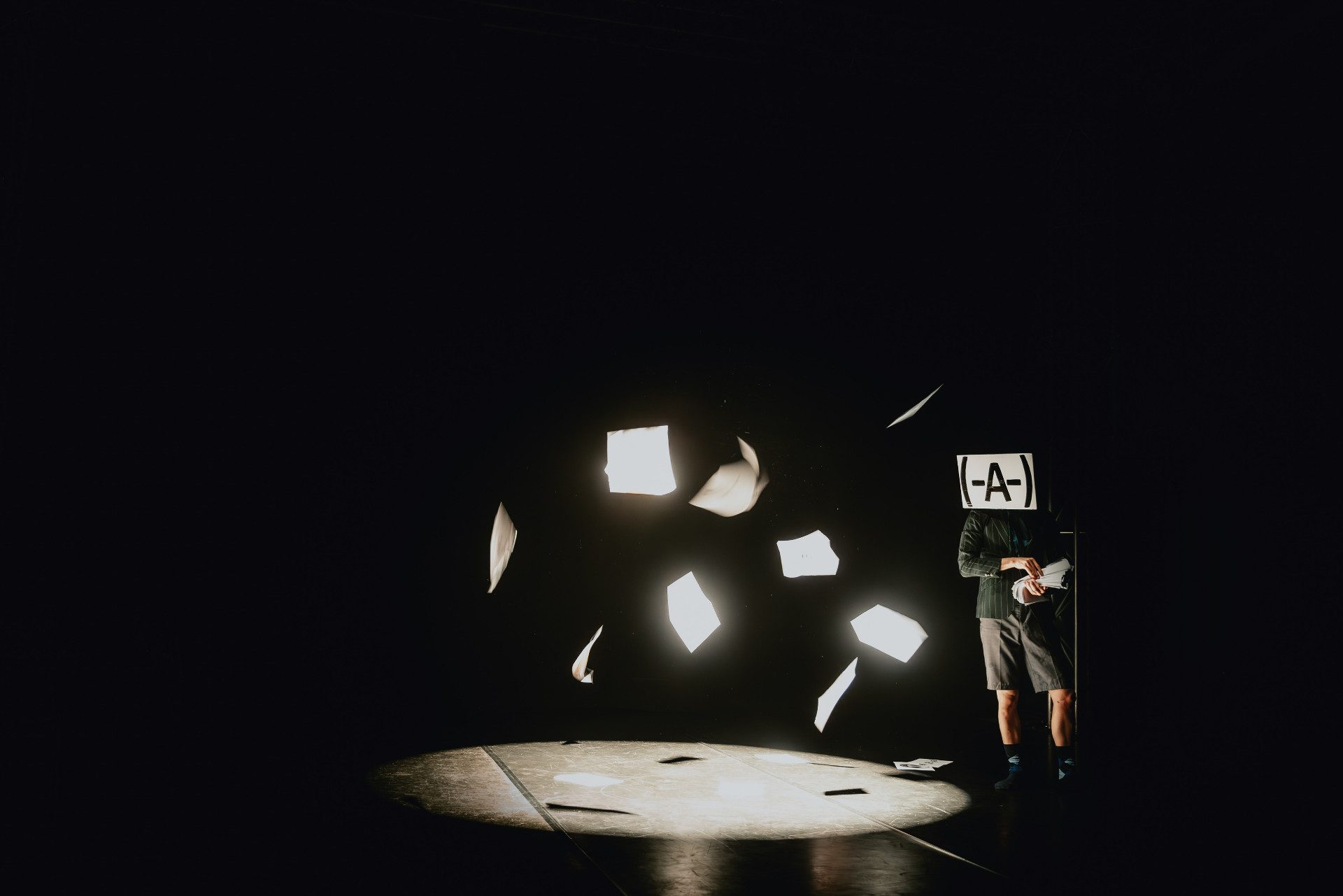 In una scena scura, un grande faro illumina il centro del palco, dove il performer lancia in aria dei fogli.
