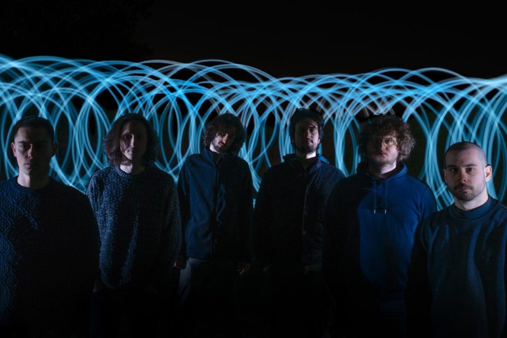 I sei componenti della band, vestiti di blu, sono circondati da luci luminose azzurre.