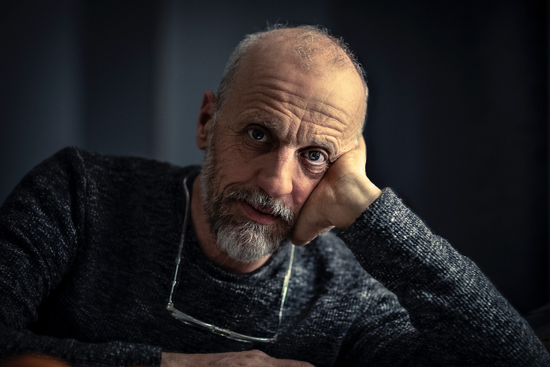 Marco Paolini. La testa appoggiata alla mano sinistra. Indossa un maglione grigio e porta gli occhiali al collo.