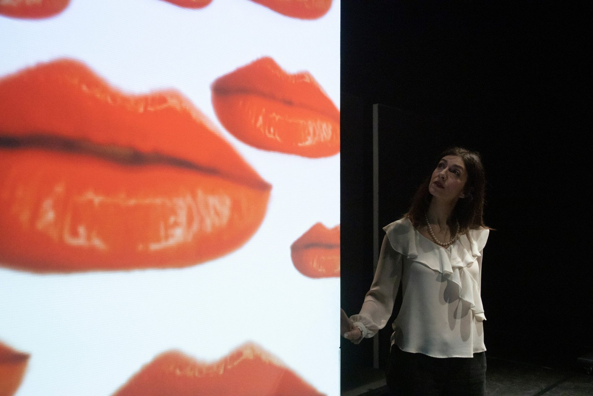 Donna con camicetta bianca e collana di perle guarda un grande schermo con immagini di labbra rosse.
