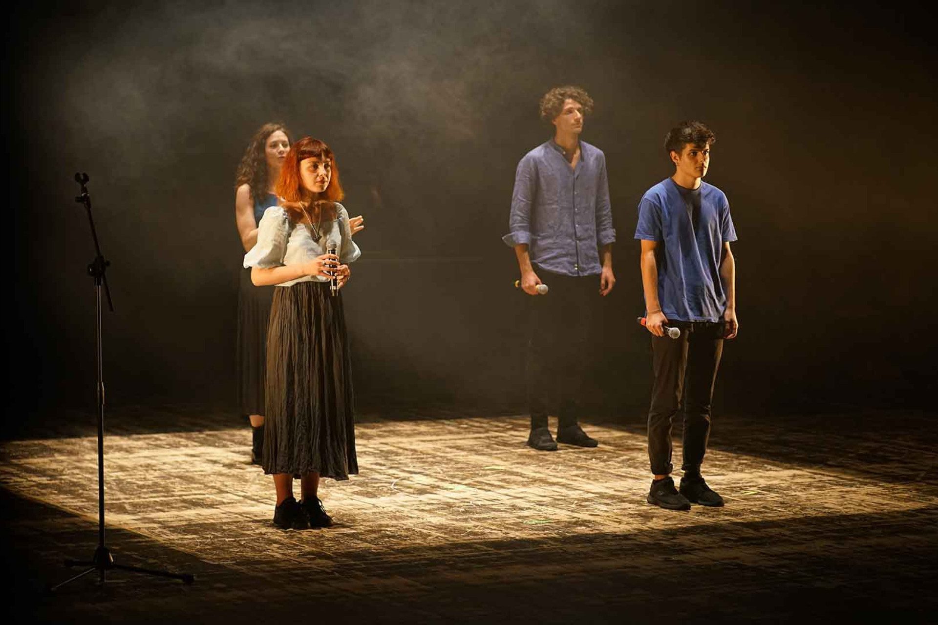 Sul palcoscenico ci sono quattro giovani attori: due ragazzi con camicia azzurra e pantaloni neri; due ragazze con gonna lunga nera e camicia. Sono disposti come le punti di un quadrato.