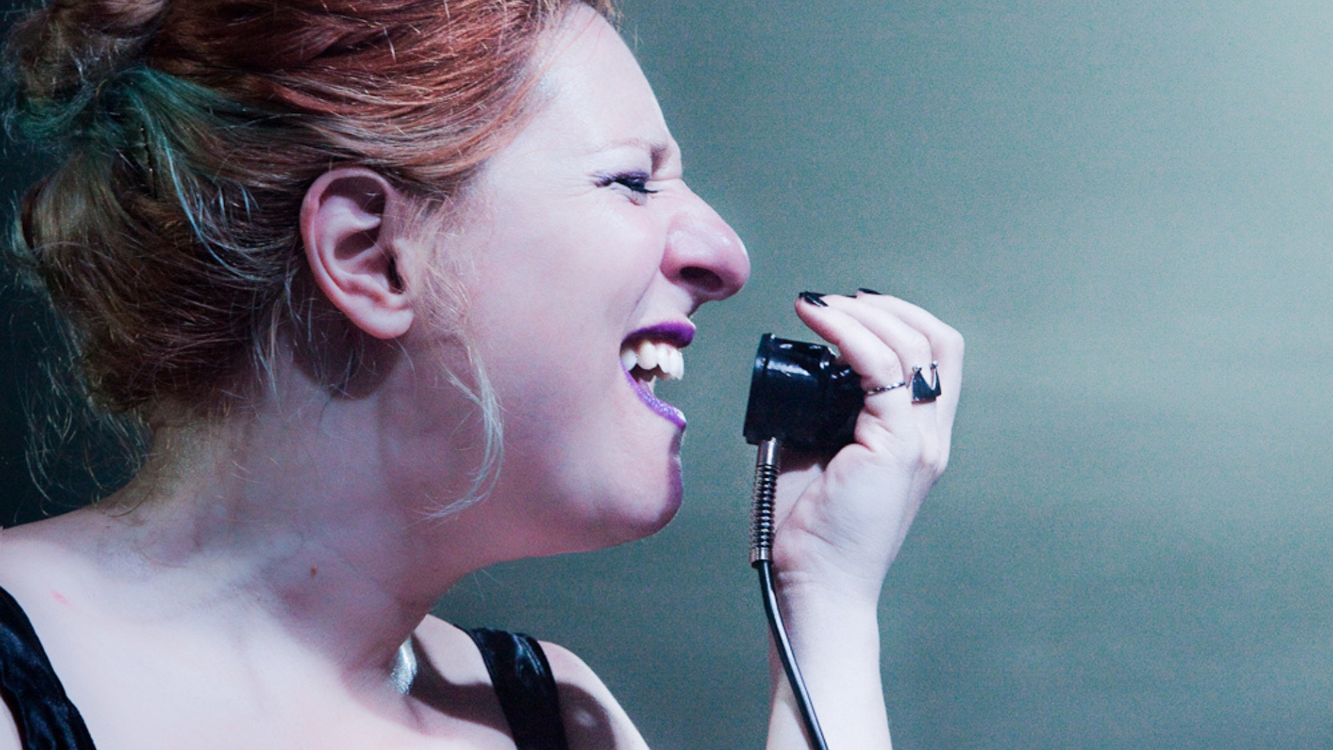 Primo piano dell'attrice Licia Lanera, in un body di pelle nera, che urla in una cornetta telefonica nera.