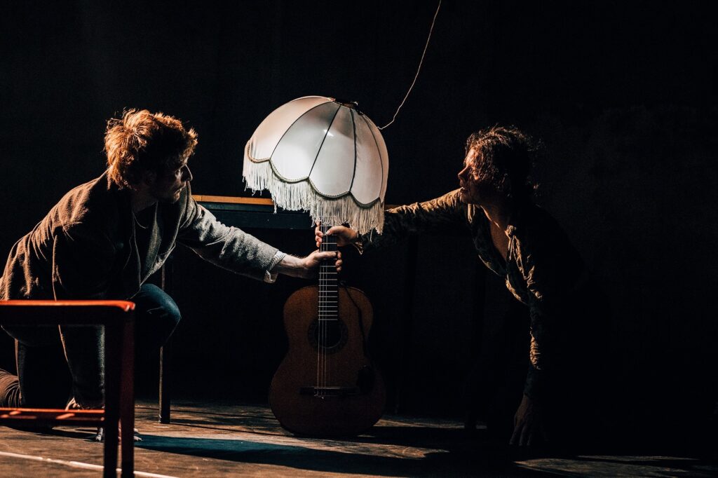 A sinistra Linus Jansner, danzatore dai capelli corti biondi, sorregge insieme a Emily Heather Manica la chitarra con in alto un paralume.